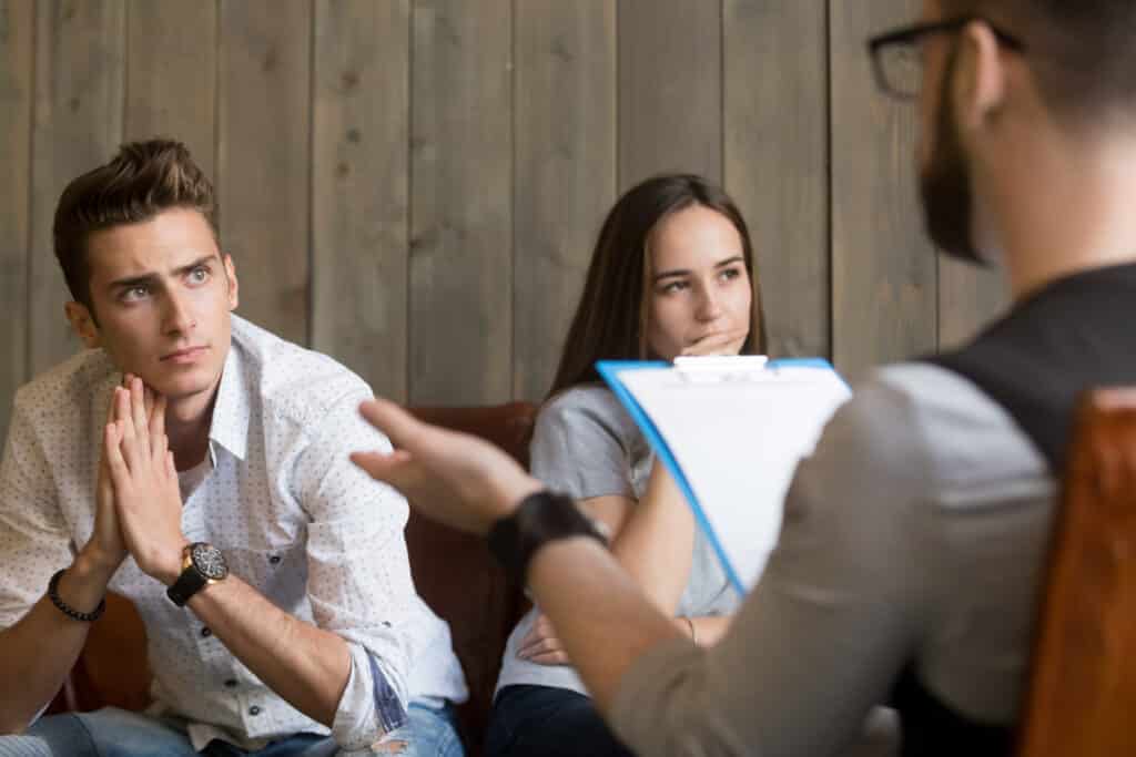 Frustrated young man listening to psychologist while his offended girlfriend sitting apart, family counselor talking to unhappy couple solving problems in relationships at counseling therapy session